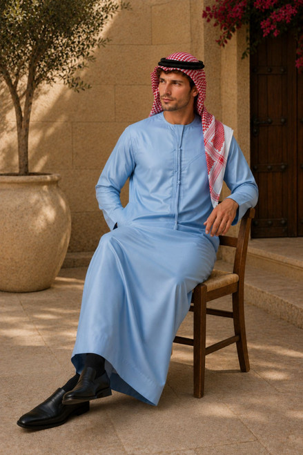 Man in traditional light blue thobe or kandora with a red and white checkered headscarf sitting on a wooden chair outdoors.