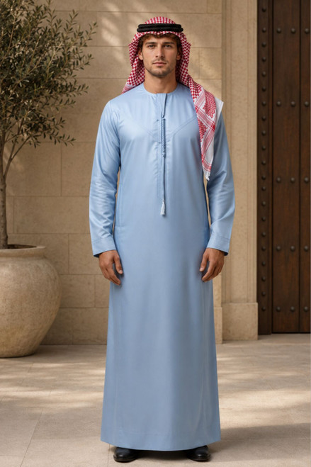 Man wearing a light blue thobe with a red and white checkered headscarf in an outdoor setting.