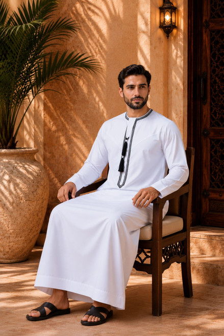 Man in traditional white thobe sitting on a wooden chair with plants and warm lighting in the background