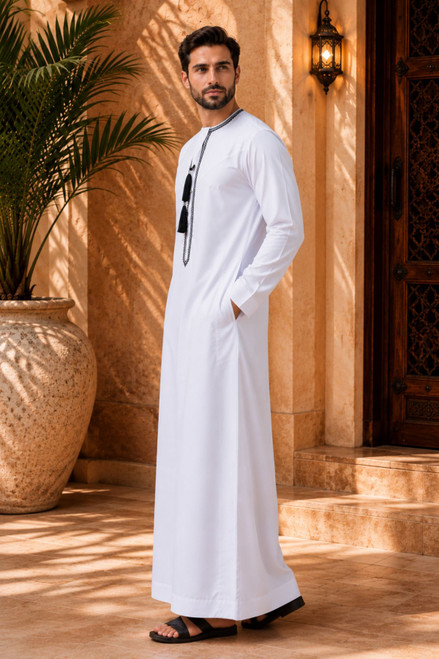 Man wearing a white thobe standing in a warm-toned indoor setting with a plant and decorative wall.