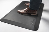 Tapis de station debout ergonomique ofinto noir en situation – gros plan avec des chaussures en cuir marron et un pantalon foncé sur le tapis antifatigue noir avec logo ofinto, pour rester debout confortablement à un bureau assis-debout
