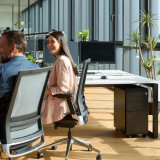 Caisson de bureau ofinto noir avec coussin d’assise dans un open space moderne – caisson mobile à tiroirs sous un bureau blanc ; une collaboratrice à un poste de travail ergonomique avec cloisons acoustiques et plantes