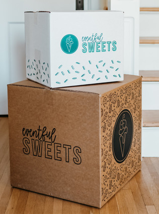 A box with a sign on it is placed on a wooden floor. The sign on the box reads ''eventful sweets''. The box itself is a storage box.  Another box nearby has a logo and sprinkles on it. The colors in the image are mostly earth tones, with accents of blue and white. The setting seems to indicate that these boxes contain sweet treats