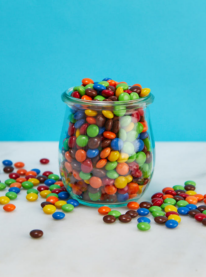 The image shows a clear glass jar filled to the brim with colorful candy-coated chocolates, likely similar to M&M's. The candies are scattered around the jar on a white surface. The background is a solid bright blue, creating a vibrant contrast with the multicolored candies. The jar contains a variety of colors, including green, red, blue, yellow, orange, and brown candies.