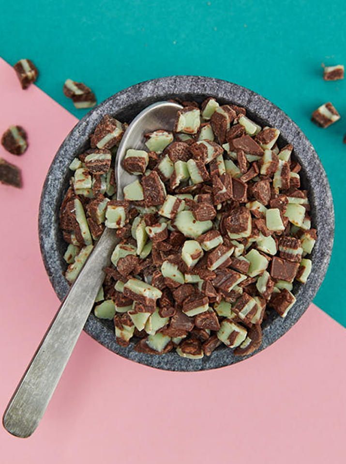 The image shows a bowl of cereal filled with chocolate and mint pieces, placed on a pink and green background. In the foreground, a spoon is partially visible.