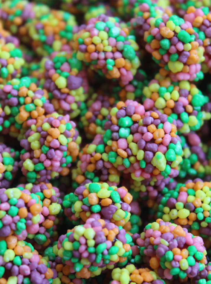 The image displays a cluster of colorful candy pieces. These candies are small and round, with a bumpy texture. The candies are covered in bright, multicolored nerds, which include vibrant shades of yellow, green, orange, pink, and purple. The overall appearance is playful and visually appealing, with a mix of colors that give the candies a festive look. The candies seem to be clustered together, almost forming a larger, irregularly shaped mass.