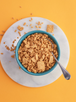 The image shows a bowl filled with crumbled cereal pieces, placed on a round white marble surface. The cereal appears to be a mixture of small, broken pieces and larger, intact squares with a textured, grid-like pattern. A metal spoon is placed in the bowl, ready to scoop up the cereal. The background is a bright orange, creating a vibrant contrast with the bowl and the marble surface. Some cereal pieces are scattered around the bowl, adding a casual and dynamic feel to the image.