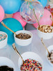 A festive scene is depicted in the image, featuring a table adorned with colorful balloons and various bowls of food. The bowls contain a variety of treats, including sprinkles, cheesecake, and other desserts. A spoon is also visible, suggesting that the food is meant to be enjoyed. The setting is reminiscent of a birthday party or celebration. The vibrant colors of the balloons and food create a cheerful and inviting atmosphere, perfect for a joyous occasion.