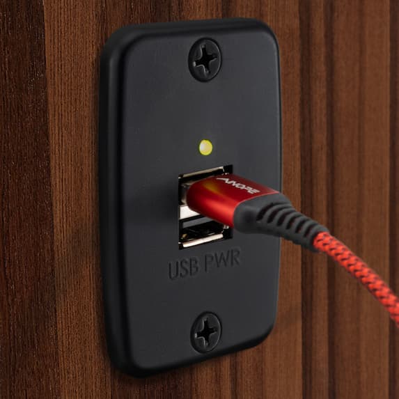 Black RV dual USB charger outlet with the indicator light on installed on a wood paneled wall with a red cord plugged in.
