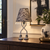 Eve lamp with a snake skin print shade and sandal feet on a dark console beneath black-and-white artwork.