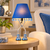 Lit blue Eve table lamp on a round bedside table in a bedroom with coordinating blue accents.