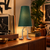 Adam green shade table lamp with silver-toned metal legs displayed on a wooden console in a warmly lit interior.
