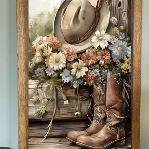 Rustic farmhouse wall sign featuring a pair of vintage cowboy boots, a weathered cowboy hat, and a colorful arrangement of wildflowers displayed on a wooden bench in a natural wood frame.