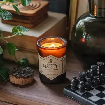 Amber glass Espresso Martini scented candle burning on a wooden table beside books, greenery, and a decorative chess set.