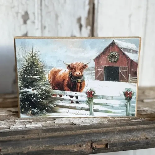 A freestanding rectangular wood block decorated with artwork of a fluffy Highland cow wearing a festive green holiday wreath around its neck. The cow stands in a snowy winter field alongside a decorated Christmas tree with a rustic red barn and evergreen trees in the backdrop under a soft pastel sky.