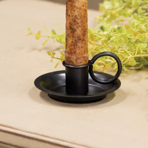 A rustic, antique-style dark iron candlestick holder designed to accommodate a standard taper candle. It features a wide dish-style base with a small, curved side handle, displaying a distressed, vintage black finish that complements farmhouse and traditional decor.