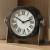 6-inch round table clock featuring a black textured resin frame, classic white dial with bold Roman numerals, and a sleek silver metal swivel stand.