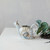 A snail-shaped stoneware watering can featuring a multi-colored white, brown, and blue reactive glaze finish, showing product dimensions of 8.75 inches in length and 6 inches in height.