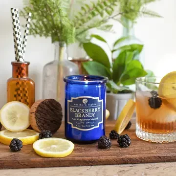 Blue glass Blackberry Brandy scented candle displayed with fresh blackberries, lemon slices, and a cocktail on a rustic wooden surface.