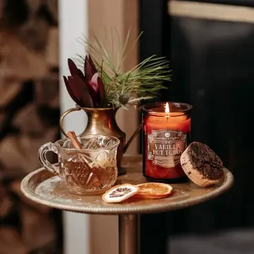 Amber glass Vanilla Hot Toddy scented candle burning beside a warm tea mug, vanilla beans, and rustic tabletop accents.