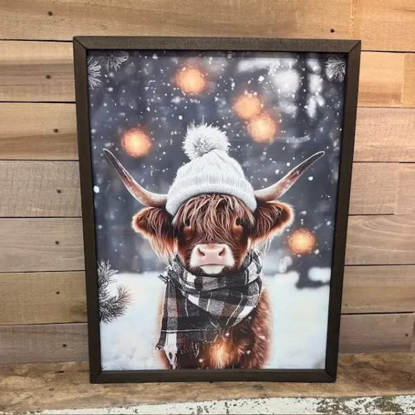 A framed art print featuring a close-up portrait of a shaggy Highland cow set against a soft, snow-dusted winter backdrop. Encased in a rustic wood frame, the artwork captures a cozy farmhouse aesthetic perfect for seasonal wall decor.