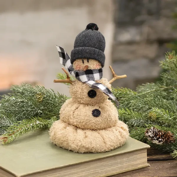 A soft plush snowman figurine designed to look like a cute, melting snow pile. It features a plush white body with coal-like black buttons, a bright orange carrot nose, black bead eyes, and a cozy knit beanie hat decorated with a festive plaid accent and pine sprig trim.