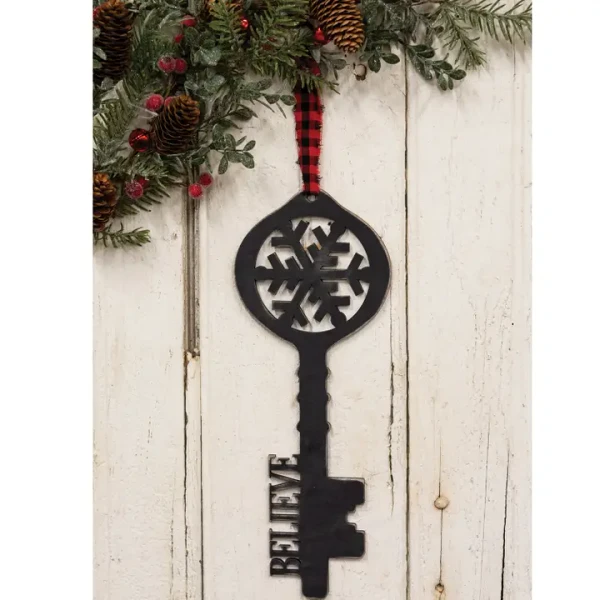 A vintage-style black wooden key decoration with a rustic weathered finish. The key features an intricate carved snowflake at the bow and the word "believe" carved along the shank. Attached at the top is a red and white gingham fabric ribbon loop for hanging on a doorknob, wreath, or Christmas tree