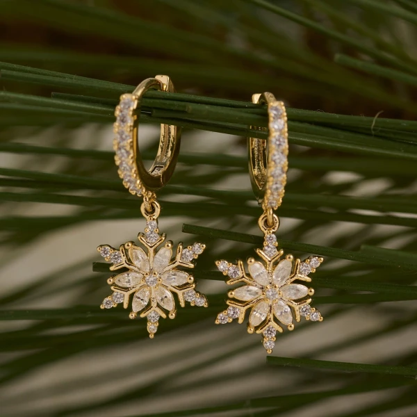 Close-up view of festive snowflake huggie hoop earrings featuring a polished hoop and dangling sparkling snowflake charm against a neutral background.