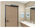 Norton Rixson 5200 Series Low Energy Power Operator installed on ADA-compliant restroom door, BSP Black Suede Powder finish, push-side arm, commercial bathroom setting with sink and mirror