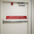 Detex ECL-395 sign installed on a commercial exit door with panic bar and door closer, showing sign placement above the push bar