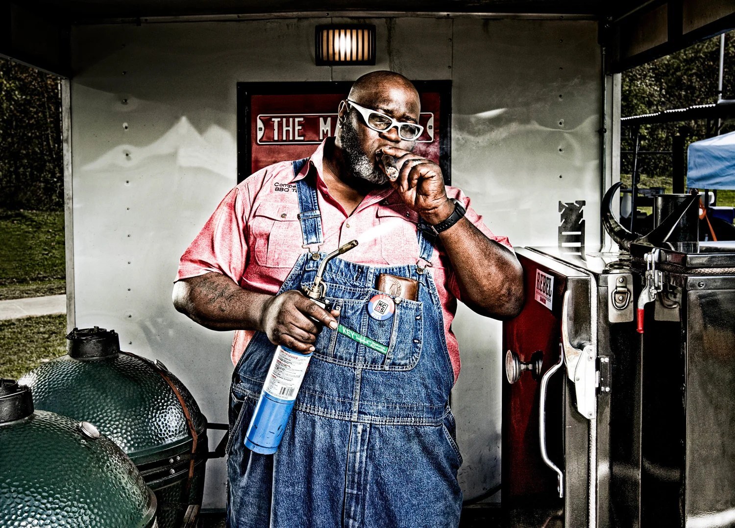 Pitmaster Moe Cason in overalls holding a torch next to kamado grills and a smoker in his competition trailer