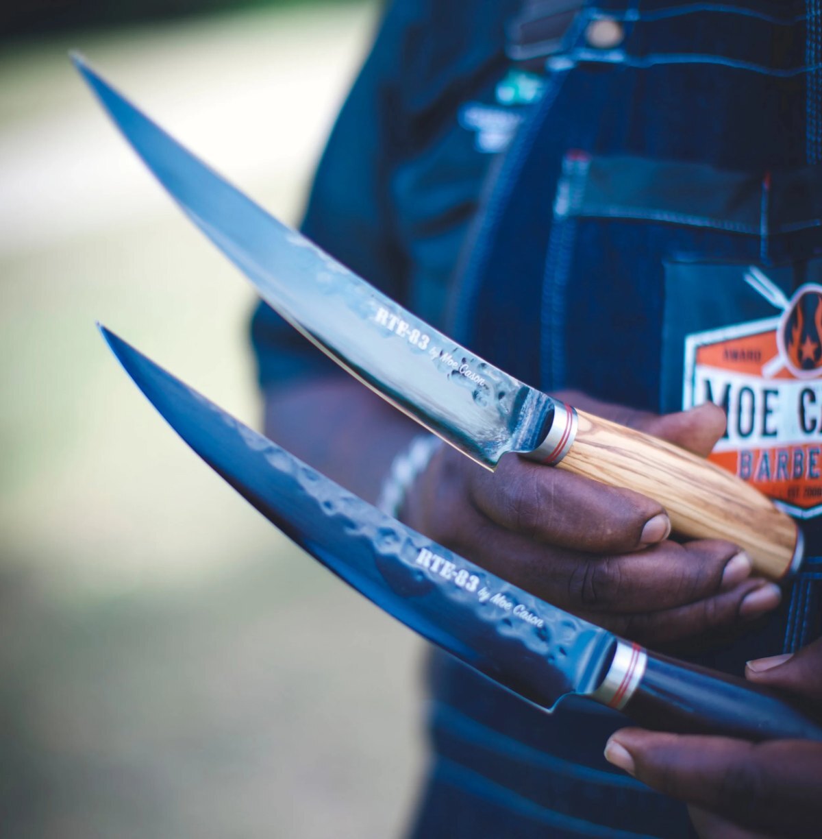 Moe Cason holding two XL boning knives with olive wood and ebony handles, engraved RTE-83 by Moe Cason