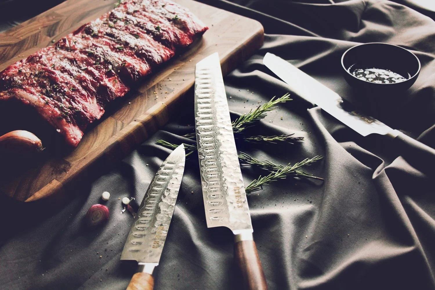 Route 83 knives on a dark cloth beside a seasoned rack of raw beef ribs on a wooden cutting board