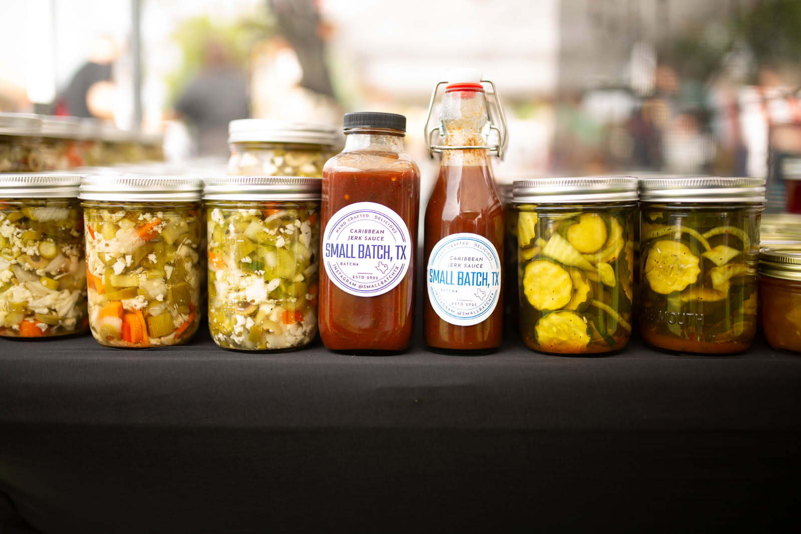 Small Batch TX giardiniera, Caribbean jerk sauce and pickle jars lined up on a farmers market table