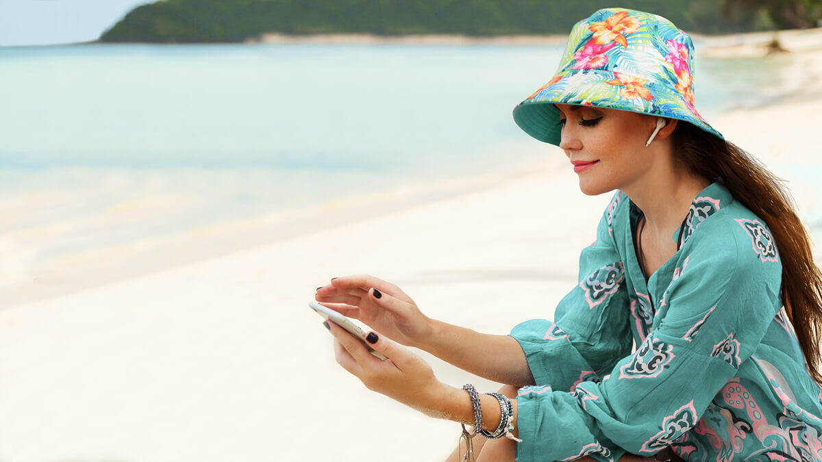 Person sitting on sandy beach near shoreline wearing colorful floral sun hat and turquoise outfit while holding smartphone with calm water in background.
