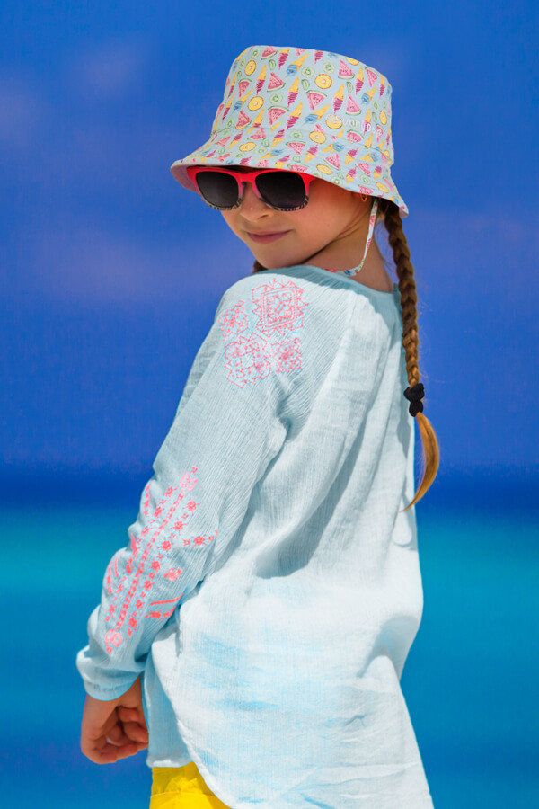 A young girl wearing a patterned bucket hat and sunglasses, standing on a beach with a blue ocean background.