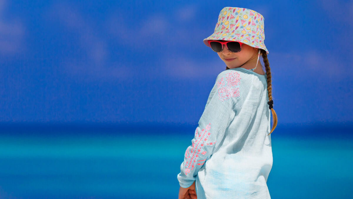 A young girl wearing a patterned bucket hat and sunglasses, standing on a beach with a blue ocean background.