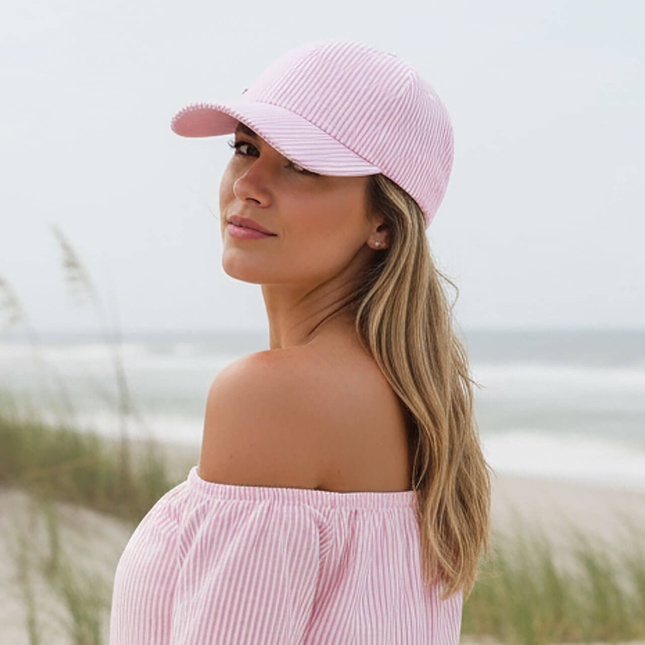 Lifestyle profile portrait of the Sun N Sand HH3593 Women’s Ball Cap in a pink and white vertical seersucker striped fabric, featuring a side metallic brand plate and a curved visor for an adjustable hook and loop fit and UPF 50+ sun protection.