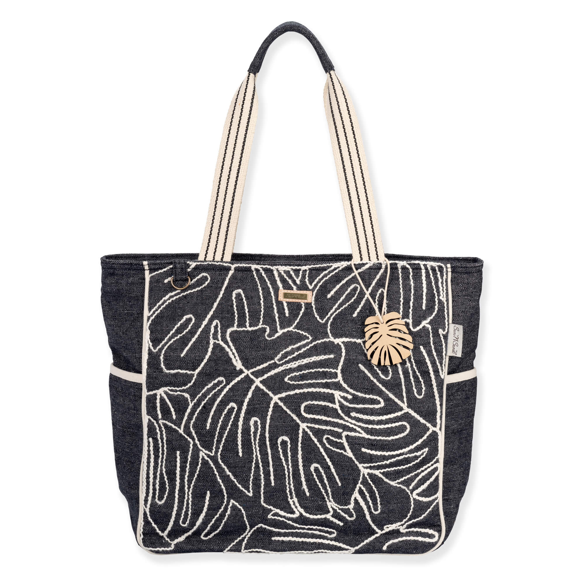 Front view of a rectangular black canvas tote featuring white embroidered monstera leaves, striped handles, and a gold leaf charm.