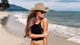 A woman with long brown hair stands on a beach wearing a tan cowboy hat, black crop top, and sunglasses.