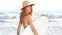 Lifestyle shot of a woman on a beach wearing a multi-colored striped fedora and holding a white surfboard.