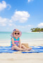 A young girl sitting on a striped towel at the beach, wearing a floral sun hat and dark sunglasses.