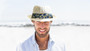 Front view of a man on a beach wearing a white perforated straw fedora with a blue tropical print band.