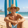 Lifestyle three-quarter view of the Sun N Sand HTT1297 men's straw lifeguard hat in Natural, featuring a blue and white striped fabric under-brim and a matching striped fabric hat band with a front logo patch for stylish sun protection and comfort during beach activities.