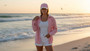 Lifestyle portrait of the Sun N Sand HH3594 Women’s Ball Cap in a soft pink cotton-blend fabric, featuring a matching pink crown and bill with a contrasting white under-bill, a small side metallic brand plate, and a curved visor for an adjustable hook