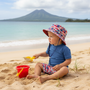 Lifestyle beach shot of the Sun N Sand HK566 infant sun hat in a red, white, and blue patriotic pattern, featuring a wide protective brim and an adjustable chin strap for reliable sun safety during beach play.