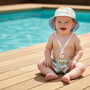 Lifestyle poolside shot of the Sun N Sand HK567 infant sun hat in a light blue submarine and sea life pattern, featuring a wide downward-sloping brim and an adjustable toggle chin strap for secure sun protection during water play.
