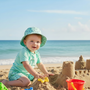 Lifestyle beach shot of the Sun N Sand HK569 infant sun hat in a mint green anchor pattern, featuring a wide protective brim and an adjustable chin strap with a toggle for secure sun safety during outdoor play.
