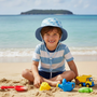 Lifestyle beach shot of the Sun N Sand HK574 Kids’ Sun Hat in a light blue palm tree print, featuring a wide downward-sloping brim and adjustable toggle chin strap for secure sun protection during beach play, outdoor activities, and seaside adventures.