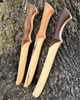 Three Elven style Daggers against the base of a moss covered tree trunk each with different grip styles. A walnut dad grip, straight cherry grip and walnut curve grip.